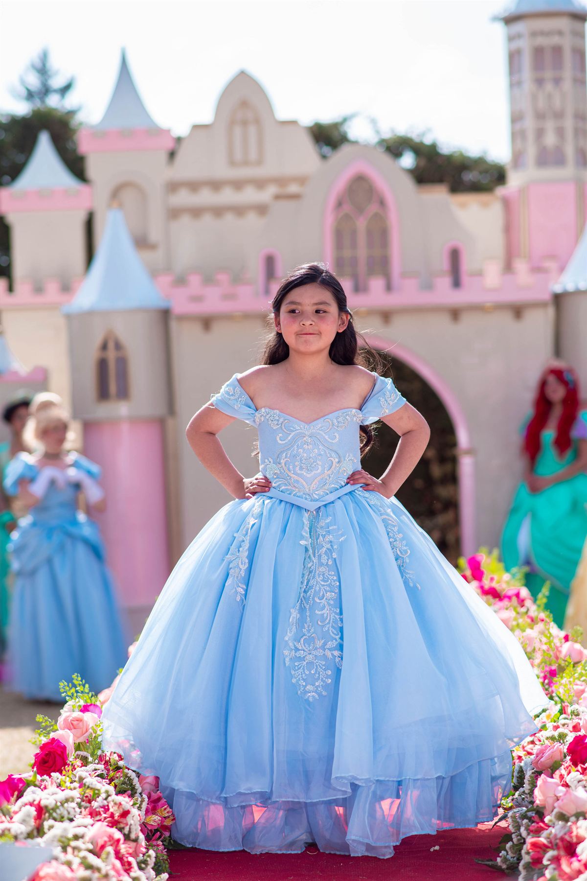 Niña con vestido celeste de falda de tul y corpiño bordado, sobre una alfombra roja entre flores